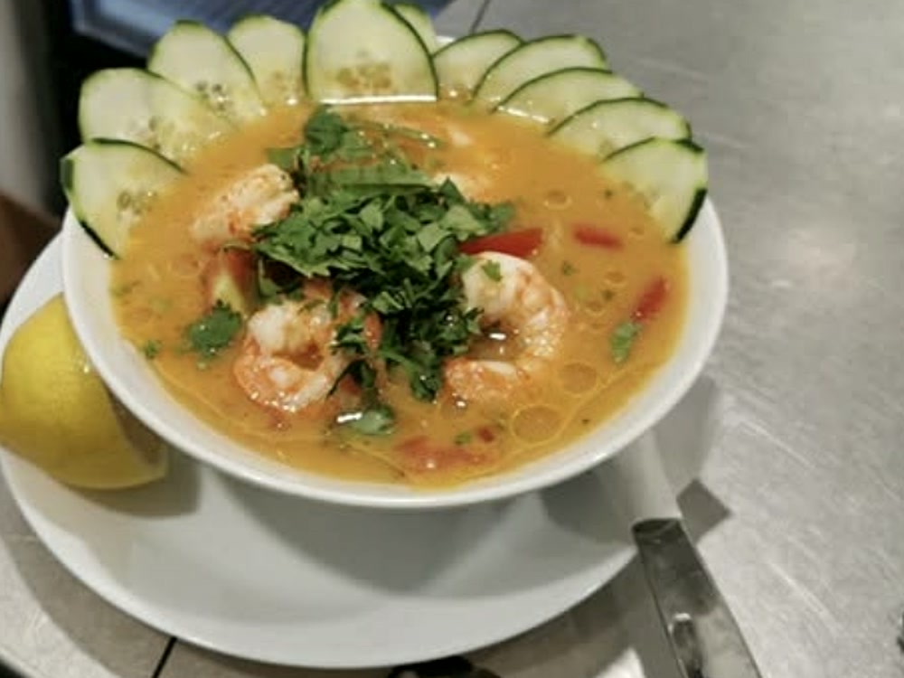Cuenco de sopa de camarones con cilantro, pepino y limón, sobre un plato blanco